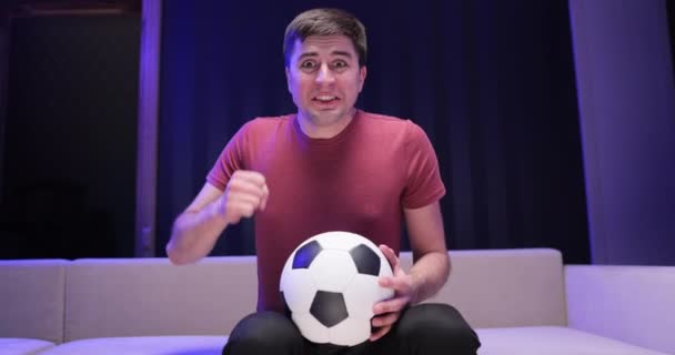 Concept de réaction des fans de sport. Fan de sport excité regardant son joueur préféré. Football fan regarder championnat tenir le ballon dans le salon sombre. Homme inquiet et remonter le moral pour le match de football à la télévision — Video