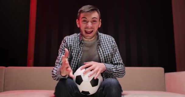 Amusant gars 30s fan de football remonter le moral soutenir l'équipe préférée tenir ballon de football en t-shirt dans le salon sombre. Les gens émotions sport loisirs style de vie concept — Video