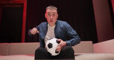 Topu elinde tutan adama yaklaş ve spor kanalını izle. Yakışıklı beyaz adam endişeleniyor ve futbol maçı için neşeleniyor. Gece geç saatte kanepede oturuyor.