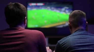 Genç arkadaşlar akşamları modern oturma odasında keyfi yerinde futbol oyunu oynayarak eğleniyorlar. Video oyunu konsepti. Adam arkadaşının maçına yüksek sesle gülüyor. Ağır çekimde çekiyoruz.