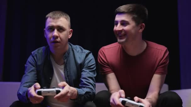 Two young guys laugh. Boys sitting on the couch in front of the TV and playing a game console, holding a joystick. Mans while gaming with a wireless controller — Stock Video