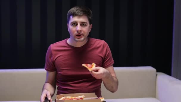 Porträt eines jungen Hipsters, der Pizza isst, sich Lieblingskomödien im Fernsehen ansieht, während er es sich auf dem Sofa im Wohnzimmer gemütlich macht. Glücklicher Mann isst italienisches Essen. — Stockvideo
