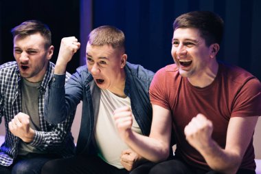 Sadık futbol taraftarları takımlarını destekliyor. Üç adam kanepede oturmuş arkadaşlarıyla futbol ya da basketbol maçı izleyip tezahürat yapıyor ve televizyon seyrediyor.