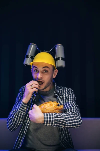 Close up of the young funny man with beer helmet on the head eating chips while watching movie on the sofa in the living room