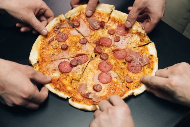 Üst düzey erkek eller pizzayı peynirli, domatesli ve jambonlu alıyor. Bir grup aç arkadaş masada oturup arka planda lezzetli öğle yemeklerini paylaşıyorlar.