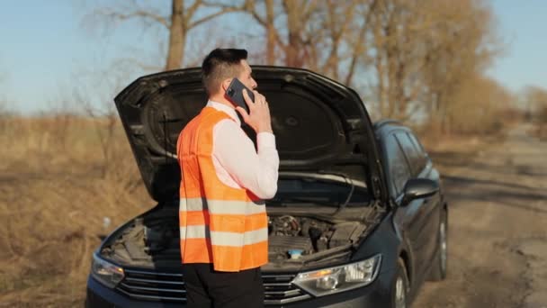 Um homem encalhado com um carro avariado pede ajuda. Jovem de pé falar por telefone chamando serviços de assistência de carro, perto do carro quebrado abriu o capô olhar reparação de ajuda rodoviária. Sinal de paragem de emergência — Vídeo de Stock