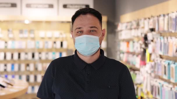 Portret van een jonge verkoper met gezichtsmasker. Knappe man elektronica winkel werknemer met medisch masker. Assistent bij gadget store — Stockvideo