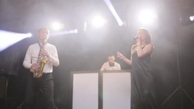 Sahnede şarkıcıyla konser veren müzik grubu. Vokalist kadın müzik grubu, saksafoncu saksafoncu, DJ adam konser müzisyeni sahnesinde ışıklar altında şarkı söylüyor.