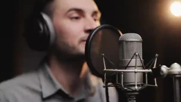 Profesyonel kayıt stüdyosunda mikrofon önünde kulaklık takan beyaz şarkıcı ışıkla şarkı kaydediyor. Müzisyenler bir şarkı üzerinde çalışıyor.. — Stok video