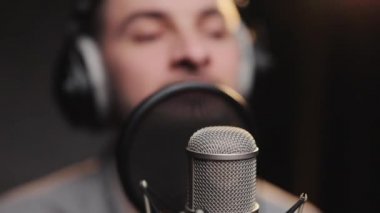 Beyaz tenli, kulaklık takan şarkıcı, düşük ışıklarla müzik stüdyosunda kayıt yaparken mikrofonla şarkı söylüyor.