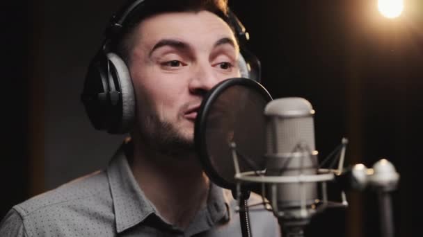Κοντινό πλάνο ενός νεαρού επαγγελματία χαμογελαστού καυκάσιου τραγουδιστή που φοράει ακουστικά εκτελεί ένα νέο τραγούδι με μικρόφωνο ενώ το ηχογραφεί σε ένα στούντιο μουσικής — Αρχείο Βίντεο