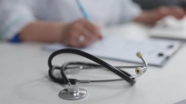 Arka planda çalışan kadın doktorla birlikte steteskop tıbbi ekipmanlarını kapatın. Masada dizüstü bilgisayar kullanarak notlar yazan profesyonel bir doktor.
