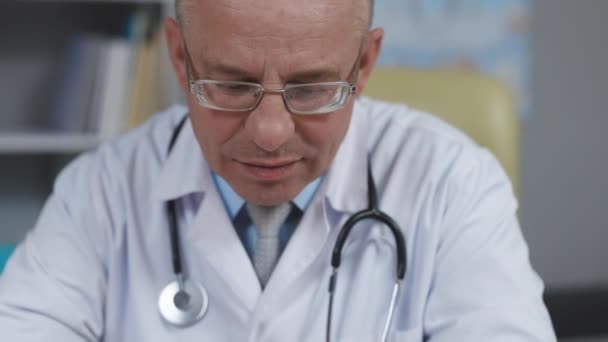 ガラスの穏やかな家族医学の医者は健康クリニックのラップトップコンピュータで働いています。ホワイト・ラボ・コートの医師が病院の机の後ろで病歴を閲覧 — ストック動画