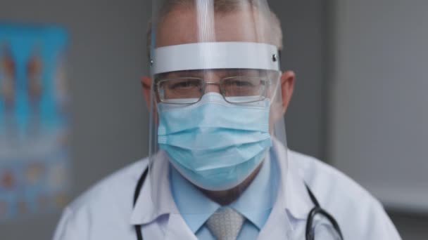 Porträtt av en manlig doktor i glasögon Bär Transparent Protective Face Shield, Mask och Overalls i ett sjukhusrum på Covid-19. Sjuksköterska i medicinsk skyddsuniform — Stockvideo