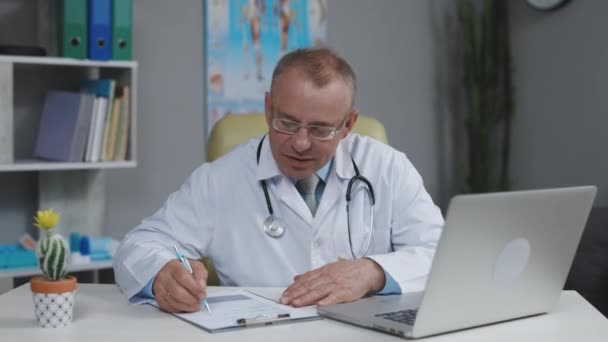 Focused middle aged senior head doctor in white medical coat and glasses sitting at workplace, talking to patient making video call on laptop, writing notes in paper journal — Stock Video