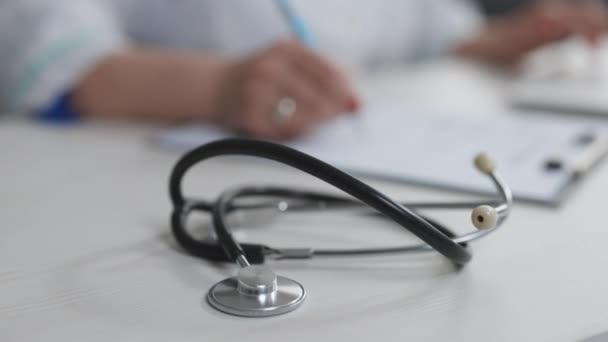 Professional physician wearing white coat writing notes using laptop at desk. Close up of stethoscope medical equipment with female doctor working in background — Stock Video