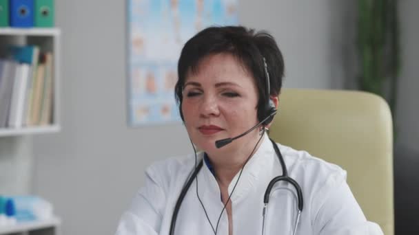 Portrét asistentky nosí bílý kabát a sluchátka se dívá do kamery a usmívá se v nemocniční kanceláři. Telemedicína, koncept vzdálených zdravotnických služeb — Stock video