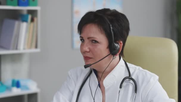 El médico exitoso en bata de laboratorio blanca usa auriculares que miran a la cámara y sonríen en la oficina del hospital. Telemedicina, telesalud. Concepto medecino moderno — Vídeo de stock