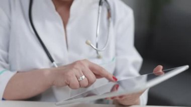 Orta yaşlı, beyaz üniformalı bir kadın doktor elinde dijital tabletle hastaları ziyaret ediyor. Olgun doktor, modern uygulama kullanarak cihazdaki sağlık geçmişini kontrol ediyor.