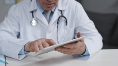 Orta yaşlı, beyaz üniformalı bir doktor elinde dijital tabletle hastaları ziyaret ediyor. Doktor, modern uygulama kullanarak cihazdaki sağlık geçmişini kontrol ediyor..