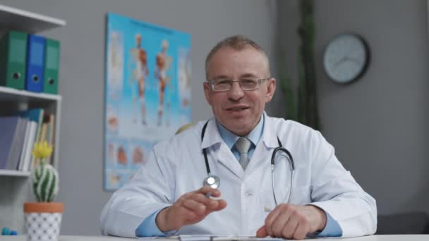 El médico de familia está sentado frente a la cámara, saludando con la mano y hablando en una clínica de salud. Médico exitoso en bata blanca de laboratorio mira a la cámara y sonríe en la oficina del hospital — Vídeos de Stock