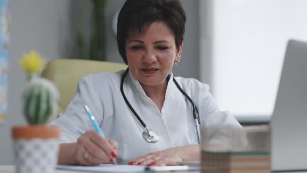 Medicul senior de vârstă mijlocie, în haină medicală albă, stând la locul de muncă, vorbind cu pacientul făcând apel video pe laptop, scriind note în jurnalul de hârtie — Videoclip de stoc