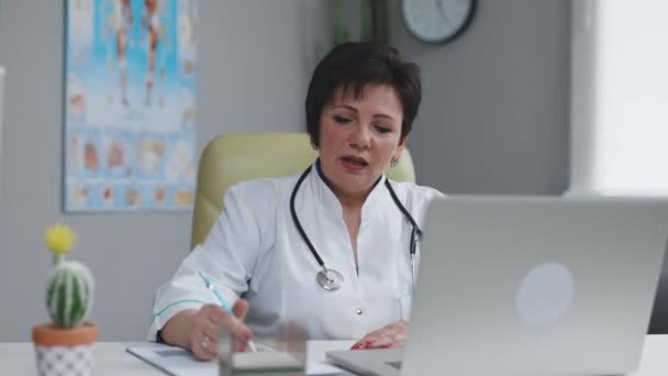 Förste chefsläkare i vit medicinsk rock sitter på arbetsplatsen, pratar med patienten gör videosamtal på bärbar dator, skriver anteckningar i pappersdagbok. Telemedicin, distansvård — Stockvideo