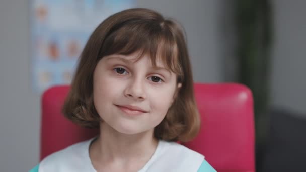 Grappig meisje dat naar de camera kijkt zit thuis op de bank. Schattig kind met mooi gezicht. Het concept van een gelukkige jeugd. Portret van meisje in medisch uniform kijkend naar camera in slow motion — Stockvideo