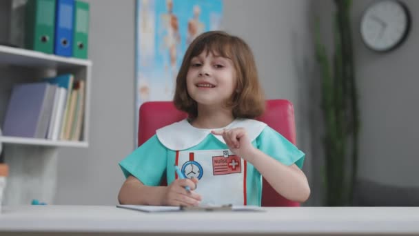Αστείο μικρό κορίτσι χαμογελώντας κοιτάζοντας κάμερα στο σπίτι, χαριτωμένο παιδί φορούν ιατρικά ρούχα μιλώντας με κάμερα κάνοντας online βιντεοκλήση ή εγγραφή vlog διασκεδάζοντας, προνήπιο παιδί με όμορφο πρόσωπο — Αρχείο Βίντεο