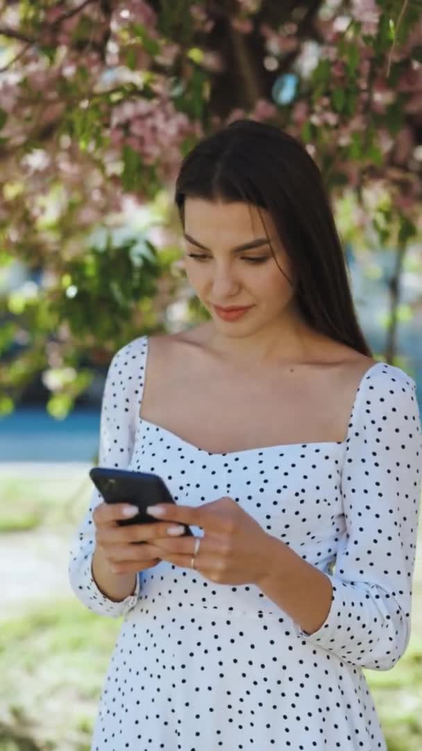 Κάθετη πορτρέτο μιας όμορφης γυναίκας που χρησιμοποιεί ένα smartphone στο πάρκο σε φόντο δέντρων σακούρα. Νεαρό όμορφο κορίτσι περιήγηση στο Διαδίκτυο χρησιμοποιώντας τα κοινωνικά δίκτυα — Αρχείο Βίντεο