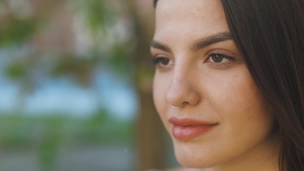 Primer plano retrato de la cara de mujer joven ojos abiertos mirando se sienten felices el sol del día está brillando y el viento sopla. Rostro de hermosa mujer suave con maquillaje natural en el bosque de flores de cerezo. — Vídeo de stock