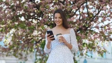 Beyaz elbiseli genç bir kadın şehir merkezinde yürüyor, akıllı telefon kullanıyor ve kahve içiyor. İletişim, çevrimiçi alışveriş, sosyal ağ kavramı