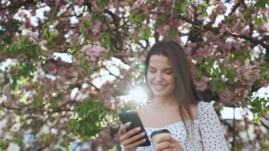 Şehir parkındaki Sakura ağacının arka planında elinde akıllı telefon ve bir fincan kahveyle çekici genç bir kadın. Beyaz elbiseli güzel bir yaz kadını cep telefonuna bakıyor ve gülümsüyor. Şık Kıyafet