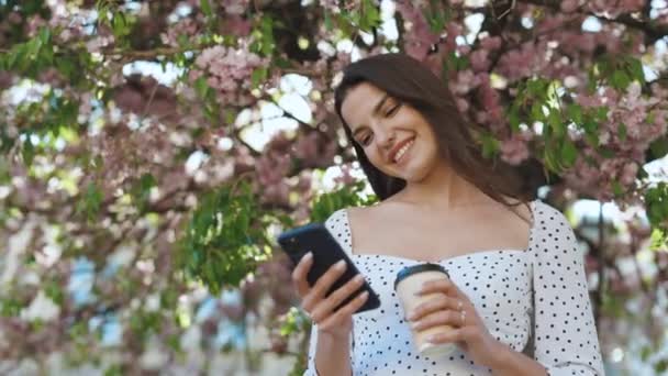 Mujer con encantadora sonrisa caminando por el parque y usando el teléfono móvil con ropa elegante. Chica usando Smartphone y bebiendo café. Comunicación, compras en línea, concepto de red social — Vídeos de Stock