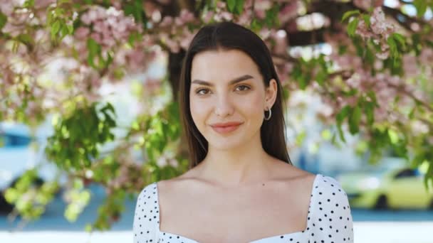 Retrato de uma mulher de cabelos escuros lindo sorrindo vestindo Sammer Vestido Charmingly enquanto em pé no parque em um fundo de árvores sakura. Jovem feliz gosta da vida. — Vídeo de Stock