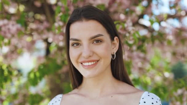 Close up portrait Smiling attractive young adult woman model looking at camera, happy beautiful 20s brunette lady pretty face dental smile posing stand alone at park on a background of sakura trees — Stock Video