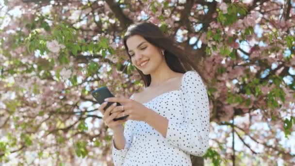 Retrato de mulher hipster feliz digitando por telefone celular ao ar livre. Menina alegre andando com smartphone em fundo urbano. Sorrindo senhora segurando celular em mãos fora — Vídeo de Stock