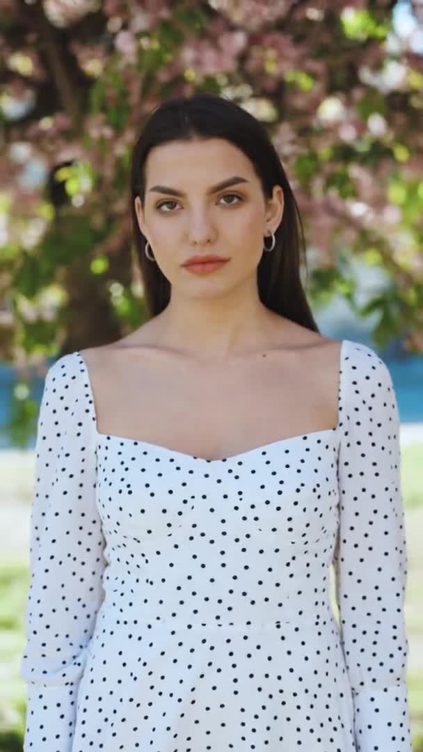 Portrait vertical de jeune femme américaine de beauté avec la coiffure longue debout sur la rue près des arbres sakura, regardant amical à la caméra. Visage humain, idéal naturel maquillage concept de peau. — Video