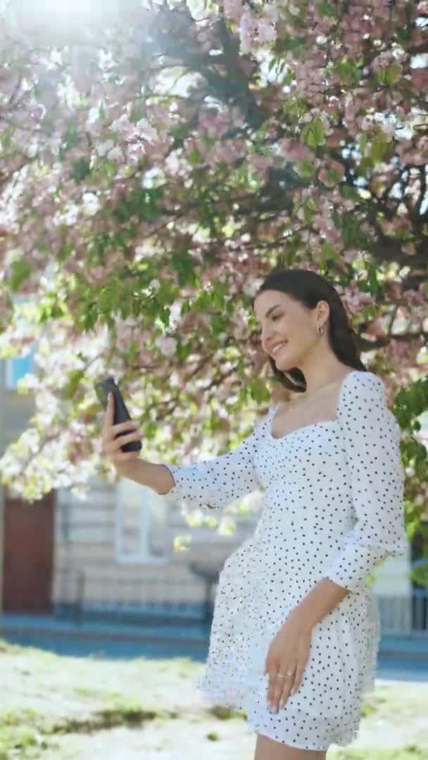 Κάθετη μορφή βίντεο από την όμορφη χαρούμενη νεαρή Καυκάσια γυναίκα με λευκό φόρεμα ποζάροντας στην κάμερα smartphone και βγάζοντας selfie φωτογραφία. Αρκετά κομψό γυναίκα τουρίστρια λήψη φωτογραφιών με κινητό τηλέφωνο — Αρχείο Βίντεο