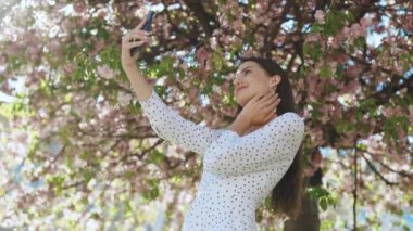 Yazlık beyaz elbiseli, gülümseyen genç bir kadın akıllı telefondan kendi portresini çekiyor. Park Sakura ağaçlarının arka planında poz vermek. Olumlu yüz duyguları gösteren bir kadın..