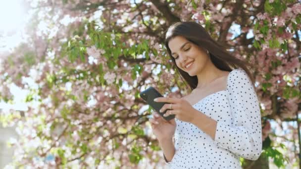 Cámara lenta Mujer alegre mirando el teléfono móvil en la calle gente urbana con el teléfono celular en la mano. Mujer de verano bonita en vestido blanco camina por la calle mirando su teléfono móvil. — Vídeos de Stock