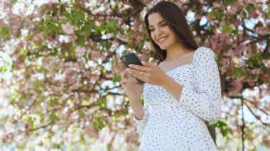 Dışarıda cep telefonuyla yazan mutlu hippi kadın. Sakura ağaçlarının arka planında akıllı telefonlu neşeli bir kız. Dışarıda cep telefonunu tutan gülümseyen kadın.