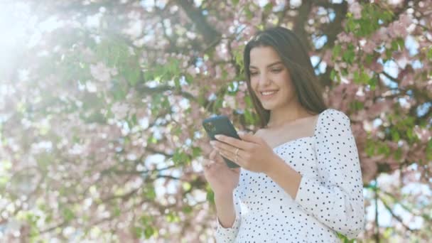 Mujer sonriente vistiendo vestido blanco usando smartphone. Chica que utiliza los mensajes de texto de aplicaciones de redes sociales reciben noticias sonriendo al aire libre. Comunicación, redes sociales, concepto de compras online. Tecnología — Vídeos de Stock