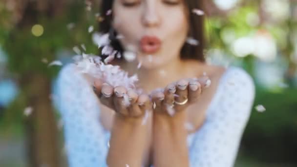 Chica soplando pétalos de flor de sakura rosa en sus manos mirando a la cámara y sonreír. Retrato hermosa chica está sosteniendo pétalos de flores de color rosa en sus manos. Pétalos que soplan, tiempo ventoso — Vídeo de stock