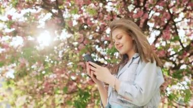 Hippi kadın dışarıda cep telefonuyla yazı yazıyor. Sakura ağaçlarının arka planında akıllı telefonlu neşeli bir kız. Dışarıda cep telefonunu tutan gülümseyen kadın..