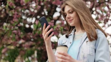 Parkta yürüyen ve şık giyinen, cep telefonu kullanan çekici gülümsemeli bir kadın. Akıllı Telefon Kullanan ve Kahve İçen Kız. İletişim, çevrimiçi alışveriş, sosyal ağ kavramı