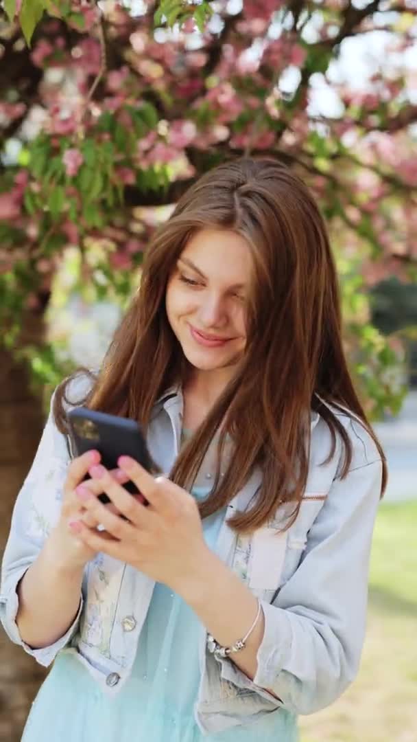 Retrato vertical de uma mulher bonita usando um smartphone no parque em um fundo de árvores sakura. Jovem menina bonita navegando na Internet usando redes sociais — Vídeo de Stock