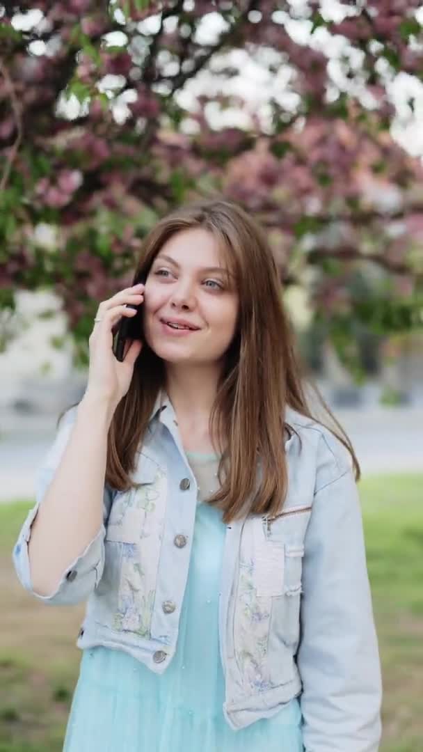 Вертикальне відео дівчина-модель розмовляє по телефону, ходячи в парку на фоні сакури. Приваблива дівчина з довгим волоссям говорить на смартфоні і посміхається на відкритому повітрі — стокове відео