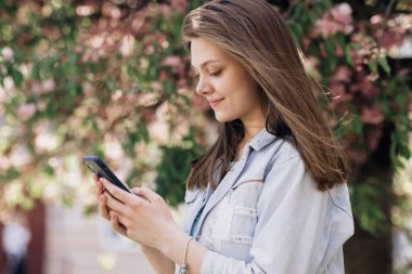 Dışarıda cep telefonuyla yazan mutlu hippi kadın. Sakura ağaçlarının arka planında akıllı telefonlu neşeli bir kız. Dışarıda cep telefonunu tutan gülümseyen kadın.