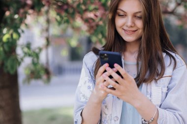 Gülümseyen kadın Central Park caddesinde yürüyor ve telefonunu kullanıyor. Beyaz elbiseli güzel bir yaz kadını cep telefonuna bakarak sokakta yürüyor.